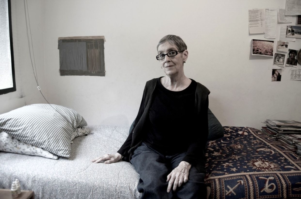 Una persona con pelo corto y canoso y gafas se sienta en el borde de una cama en una habitación amueblada de forma sencilla con fotos y papeles en la pared. La cama tiene ropa de cama a rayas y estampada. La persona mira hacia la cámara.