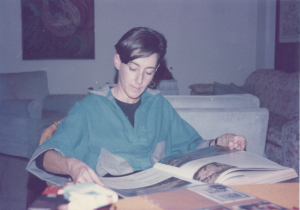 Una persona con el pelo corto está sentada a la mesa de un salón, leyendo un gran libro abierto. Lleva una camisa verde azulado y gris. El fondo muestra un sofá, un cuadro y algunos objetos dispersos sobre la mesa.