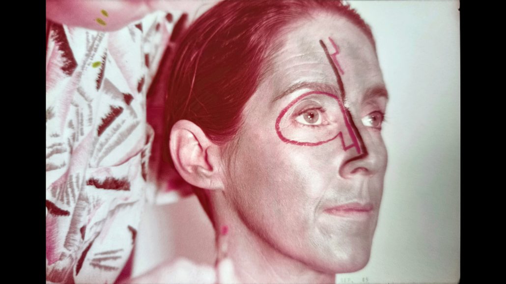 Una persona de piel clara y pelo castaño mira hacia la derecha. En su cara hay dibujada una línea roja que rodea un ojo y baja por la nariz. El fondo muestra un cojín estampado.