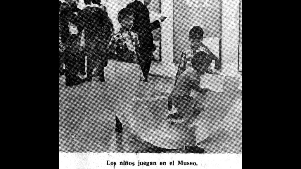 Tres niños juegan dentro de una estructura translúcida y curva en la galería de un museo. En las paredes del fondo se ven obras de arte enmarcadas. La leyenda en español dice: "Los niños juegan en el Museo.