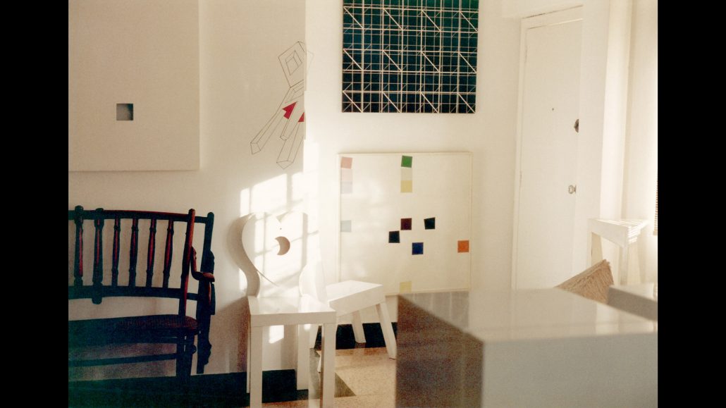 Una habitación muy iluminada con arte abstracto moderno sobre paredes blancas, un banco de madera oscura, una silla blanca y adornos geométricos en la pared. La luz del sol entra por una ventana y proyecta sombras sobre el suelo y los muebles.