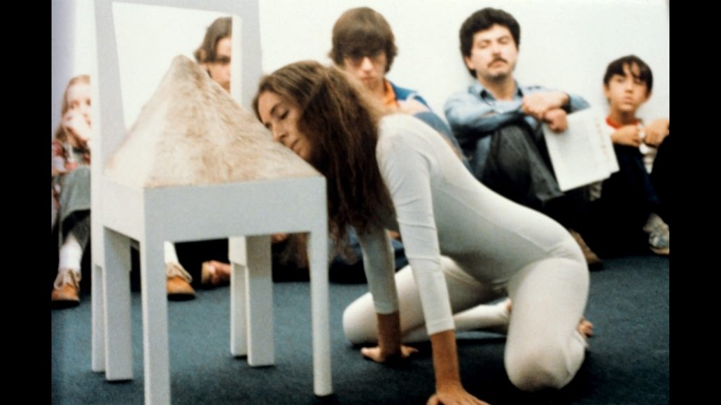 Una mujer con un body blanco se arrodilla en el suelo, apoyando la cara en el asiento de una silla cubierta de un material peludo. Varias personas sentadas en el suelo, al fondo, la observan.