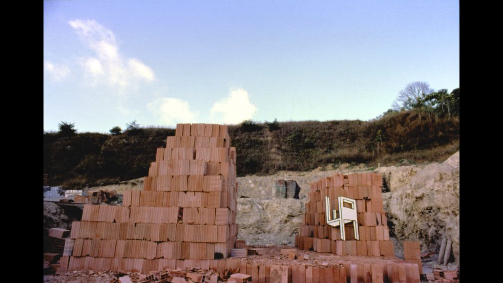 Montones de ladrillos rojizos se apilan de forma irregular en una obra, con un único marco de ventana blanco incrustado en uno de los montones. Una colina de tierra y escasa vegetación son visibles en el fondo bajo un cielo azul.