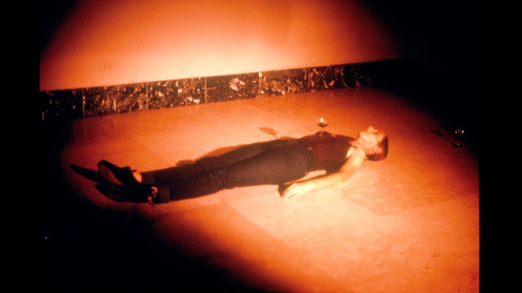 Una persona está tumbada boca arriba sobre un suelo de baldosas en una habitación poco iluminada con luz rojiza. Dos copas de vino están colocadas cerca de sus manos, y la pared de detrás tiene un zócalo oscuro.