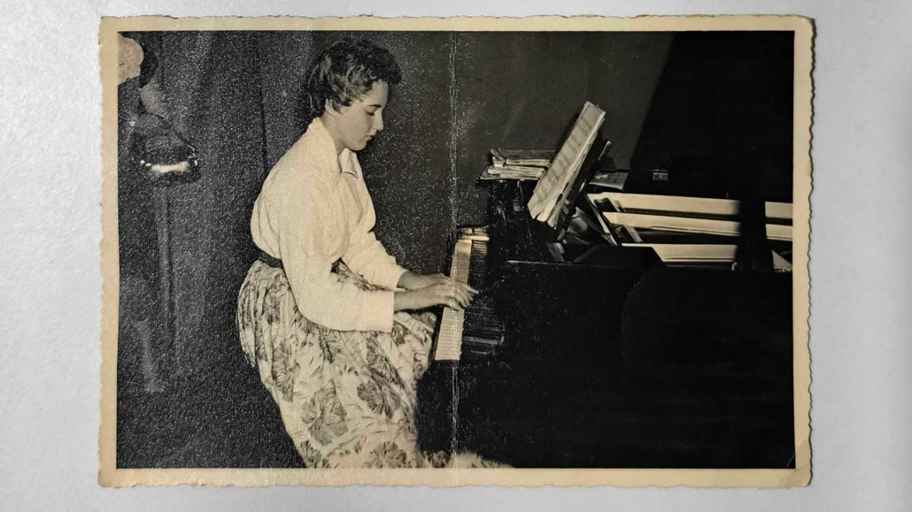 Una mujer con blusa blanca y falda estampada está sentada al piano, tocando mientras mira una partitura. La foto es en blanco y negro con los bordes ligeramente desgastados, lo que sugiere que es antigua.