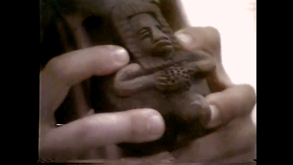 Primer plano de dos manos que sostienen una figurita de color oscuro que representa una figura sentada con un detallado tocado y sosteniendo un objeto en el regazo.