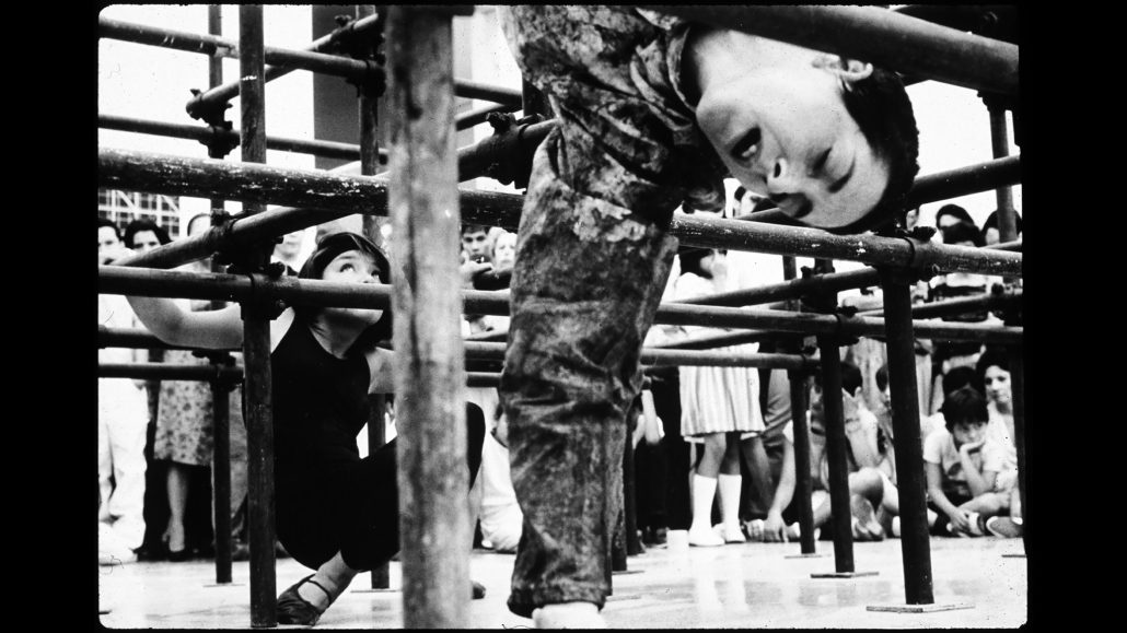 Un niño se inclina hacia atrás sobre unas barras metálicas mientras otro se agacha cerca. Al fondo, una multitud observa la actividad. La foto es en blanco y negro.