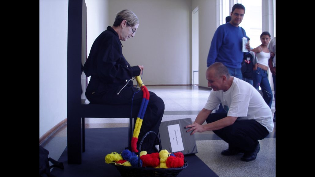 Una persona mayor se sienta en una silla de gran tamaño, tejiendo con grandes agujas e hilo grueso en rojo, amarillo y azul. Un hombre se arrodilla con un documento en la mano. En el suelo hay una cesta con hilos; al fondo, varias personas de pie.