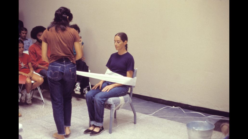 Una mujer se sienta en una silla con el torso pegado al respaldo. Otra mujer, de pie frente a ella, aplica la cinta. Hay gente sentada a su alrededor, observando la escena. Hay un cubo en el suelo.