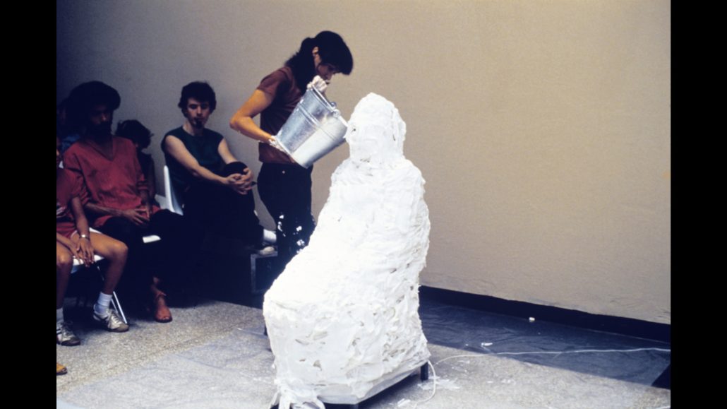 Una persona vierte yeso blanco sobre otra sentada en una silla, que ya está cubierta en su mayor parte. Varias personas sentadas observan la escena contra una pared lisa en una habitación muy iluminada.