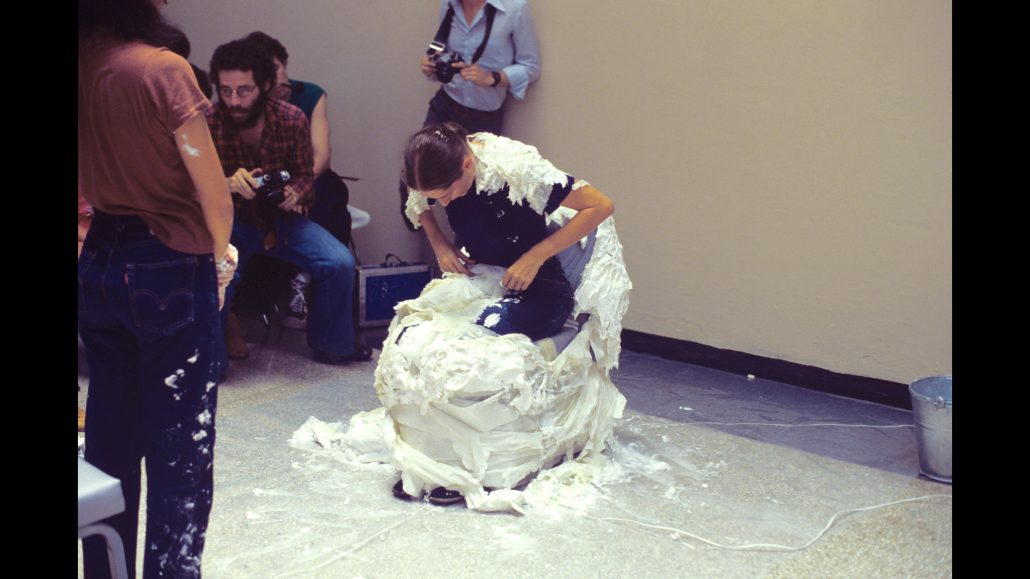 Una persona se sienta en una silla cubierta de espuma o crema blanca, con la sustancia también sobre su cuerpo. Varias personas observan a su alrededor, algunas con cámaras. La escena parece desarrollarse en un interior.