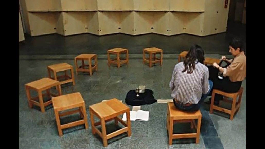 Dos personas se sientan en taburetes de madera mirando hacia otro lado, con diez taburetes vacíos dispuestos en semicírculo a su alrededor. Un libro abierto, una taza y una chaqueta están en el suelo, en el centro. De fondo, una pared divisoria de color beige.