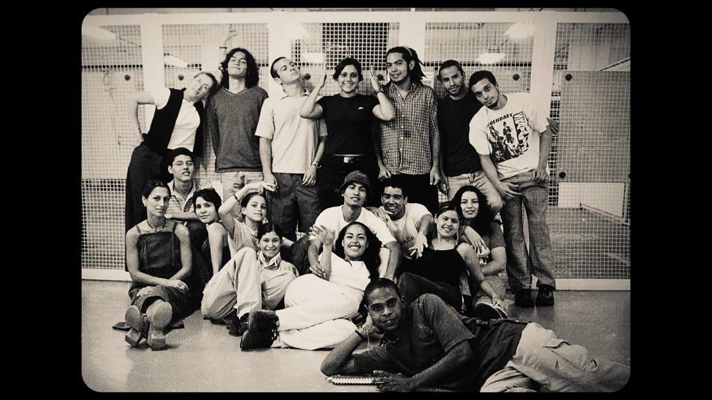 Un grupo de veinte jóvenes adultos posan juntos en un interior. Algunos están de pie, otros sentados o tumbados en el suelo, y la mayoría sonríe. El fondo muestra una alambrada y una iluminación brillante. La imagen es en blanco y negro.