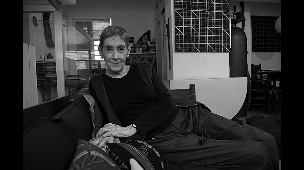 Una persona de pelo corto con camisa de manga larga y tirantes se reclina en un sofá en una habitación con estanterías, obras de arte y diversos objetos de fondo. La imagen es en blanco y negro.