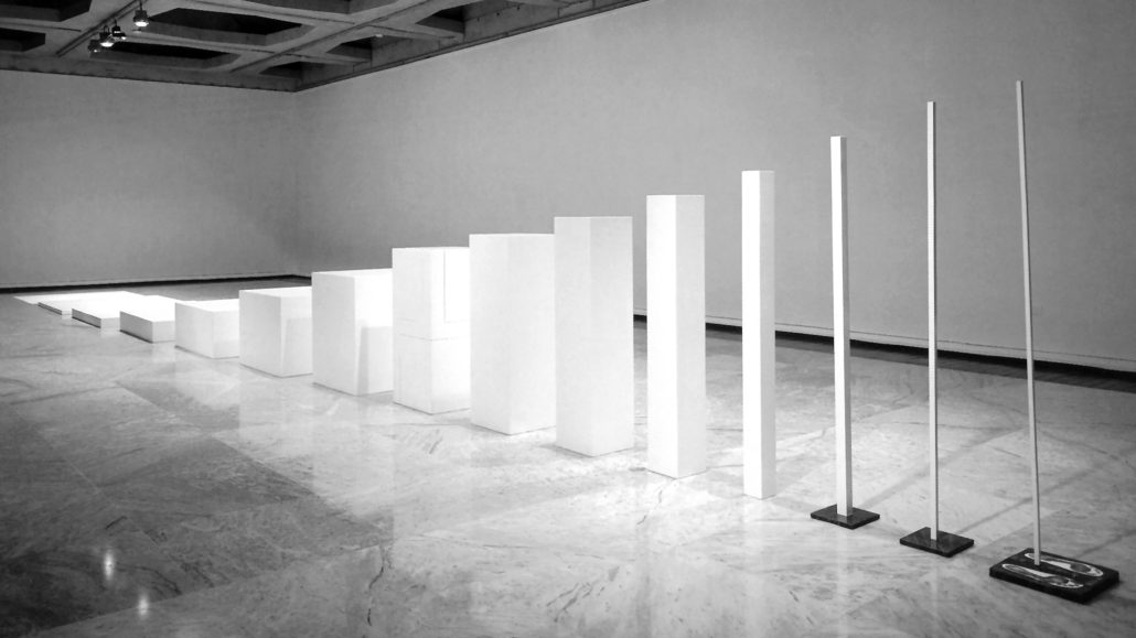 Una hilera de columnas blancas rectangulares y cuadradas de diferentes alturas y anchuras está dispuesta en una galería de arte minimalista con suelo de mármol y una pared blanca lisa. Al final de la fila hay dos postes altos y delgados.