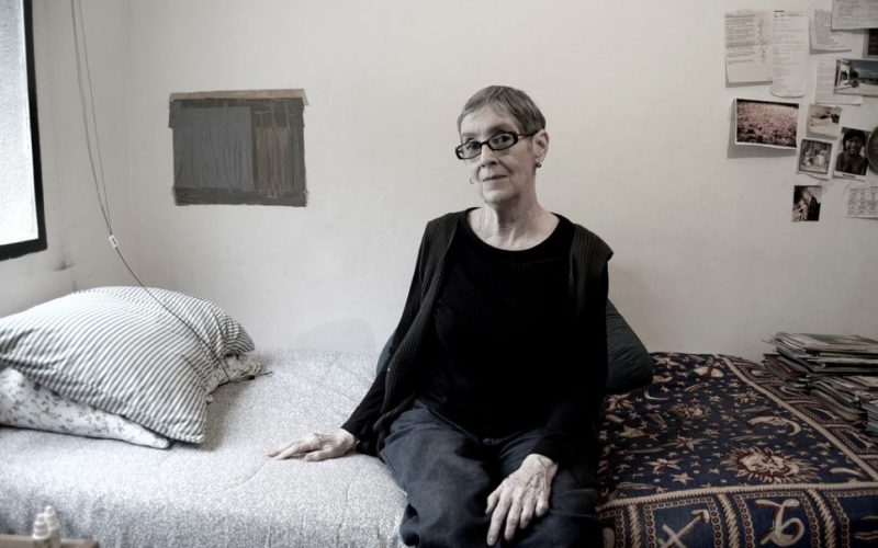 Una persona con pelo corto y canoso y gafas se sienta en el borde de una cama en una habitación amueblada de forma sencilla con fotos y papeles en la pared. La cama tiene ropa de cama a rayas y estampada. La persona mira hacia la cámara.